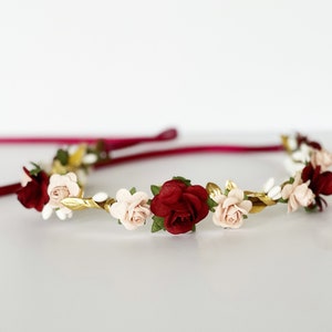 Op de afbeelding: Een bloemenkrans met donkerrode en lichtroze roosjes, afgewisseld met kleine witte bloemetjes en metallic gouden blaadjes. Het geheel is bevestigd aan een dun, donker magenta satijnen lint dat aan beide kanten van de bloemenband uitsteekt.