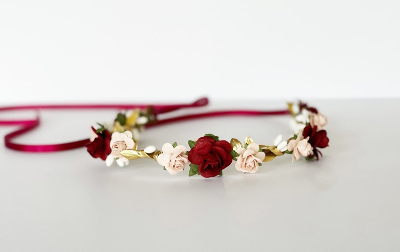 Op de afbeelding: Een bloemenkrans met donkerrode en lichtroze roosjes, afgewisseld met kleine witte bloemetjes en metallic gouden blaadjes. Het geheel is bevestigd aan een dun, donker magenta satijnen lint dat aan beide kanten van de bloemenband uitsteekt.
