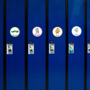 Peut inclure: Rangée de casiers d'école bleus avec des étiquettes nominatives rondes. Les étiquettes présentent des illustrations de dessins animés d'une chenille, d'un éléphant, d'une créature et d'un oiseau, chacune avec un nom en dessous. Les casiers ont des cadenas à combinaison.