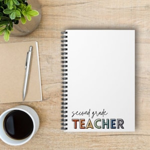May include: A white spiral notebook with the text "second grade TEACHER" in colourful letters. The notebook is on a wooden surface with a pen and a cup of coffee.