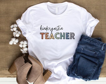 May include: White t-shirt with the text "kindergarten TEACHER" in rainbow colors. The shirt is styled with jeans and brown boots.