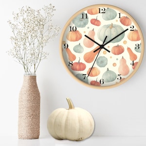 Op de afbeelding: Een ronde wandklok met een licht houten frame. De wijzerplaat heeft een crèmekleurige achtergrond met aquarel pompoenen in tinten oranje, groen en wit. Zwarte wijzers en cijfers maken het ontwerp compleet. Een witte pompoen en vaas met bloemen staan op de voorgrond.
