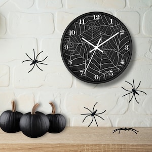 May include: Black and white clock with a spiderweb design on a white brick wall with black pumpkins and spiders. The clock face reads 12, 1, 2, 3, 4, 5, 6, 7, 8, 9, 10, 11.