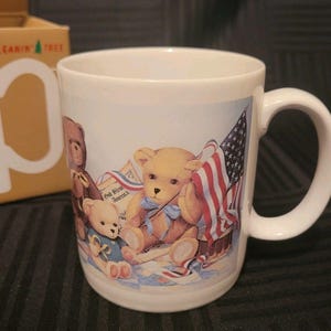 Op de afbeelding: Witte keramische mok met een patriottisch teddybeerontwerp. De mok toont een lichtbruine teddybeer met een Amerikaanse vlag en een blauwe strik. De woorden "God Bless America" staan op de mok.