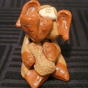 Puede incluir: Figurita de cerámica artesanal de un perro con grandes orejas y un objeto en forma de cacahuete en sus patas. El perro es principalmente marrón con rayas y detalles en beige. Estilo artístico popular.