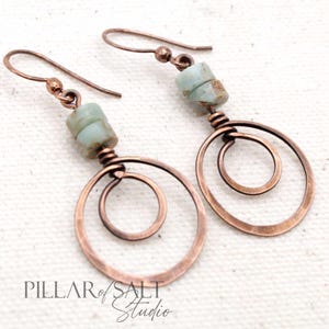 May include: A pair of copper wire earrings with a light blue gemstone bead on each earring. The earrings feature two copper wire circles, one inside the other.