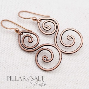 May include: A pair of copper-colored spiral earrings. Each earring features three connected swirls, with a hook for the ear. The earrings are displayed on a white surface. The earrings are from Pillar of Salt Studio.