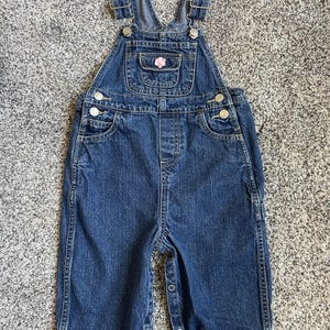 May include: Blue denim overalls with adjustable straps and snap closures. Features a small pocket with a pink flower applique. The overalls have a classic design, suitable for children.