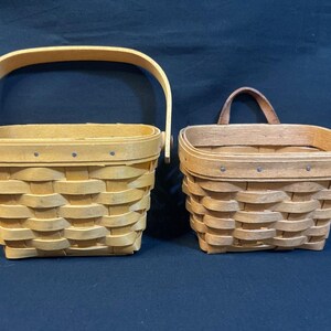 May include: Two woven baskets. The larger basket has a tall, arched handle. The smaller basket has a short, fixed handle. Both baskets are made of light-colored wood and have a rectangular shape.