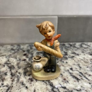 May include: A ceramic figurine of a boy holding a loaf of bread and a small jug. The boy is wearing a red scarf and has brown hair. The figurine is painted in shades of brown, red, and white.