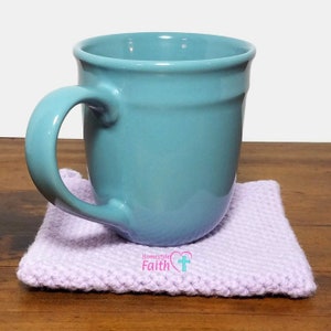 May include: A light blue ceramic mug sits on a lavender crocheted coaster. The coaster has the text "Homestyle Faith" and a pink heart with a white cross.