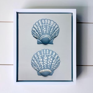 May include: A framed wall art piece featuring two blue scallop shells mounted on a light grey background. Each shell is decorated with intricate white botanical patterns and fine line details. The entire display is enclosed in a white frame.