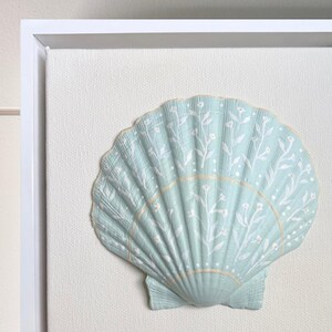 May include: A light mint green seashell mounted on a textured off-white canvas within a white frame. The shell features a hand-painted white floral vine pattern with delicate leaves and small blossoms, accented by a thin horizontal gold line.