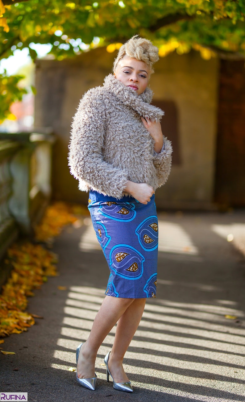 May include: A woman wearing a light brown fuzzy jacket, a blue and brown patterned skirt, and silver heels. She is standing on a sidewalk with fall leaves on the ground.