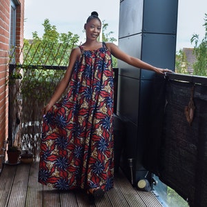 Pu&ograve; includere: Una donna indossa un lungo abito maxi fluido con un motivo stampato africano colorato. L'abito ha una vestibilit&agrave; ampia e comoda e presenta una scollatura rotonda e spalline sottili. La donna &egrave; in piedi su un ponte di legno e sorride alla telecamera.