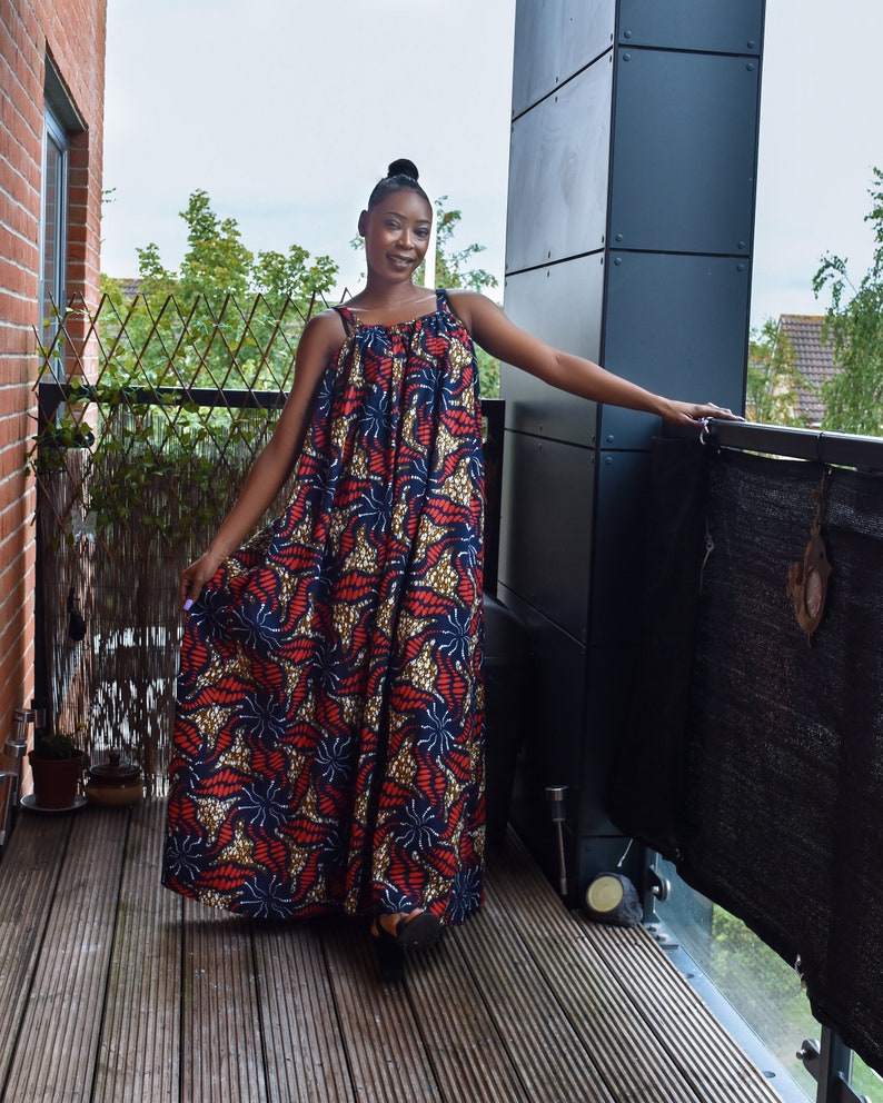 Pu&ograve; includere: Una donna indossa un lungo abito maxi fluido con un motivo stampato africano colorato. L'abito ha una vestibilit&agrave; ampia e comoda e presenta una scollatura rotonda e spalline sottili. La donna &egrave; in piedi su un ponte di legno e sorride alla telecamera.