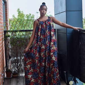 Pu&ograve; includere: Una donna indossa un lungo abito maxi senza maniche con una stampa astratta colorata. L'abito ha una scollatura arricciata e una silhouette ampia e fluida. La donna &egrave; in piedi su un ponte di legno con una ringhiera di metallo dietro di lei.