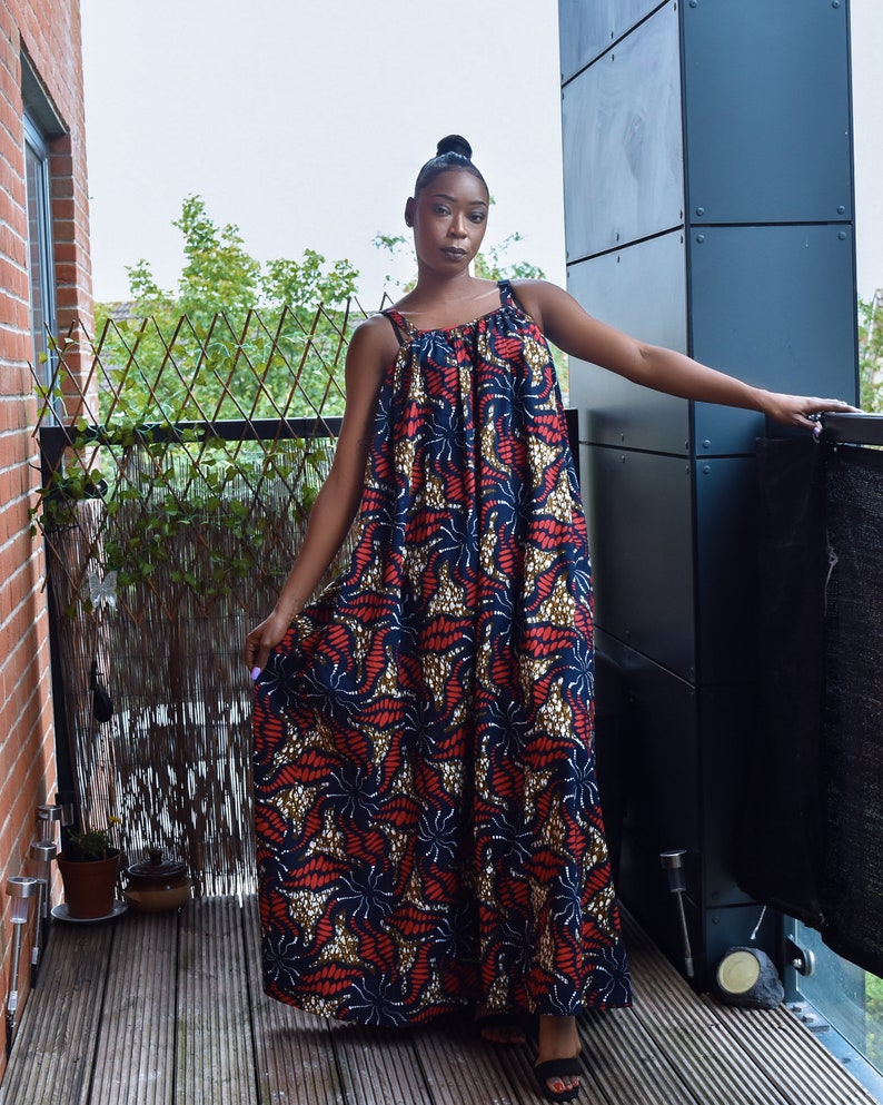 Pu&ograve; includere: Una donna indossa un lungo abito maxi senza maniche con una stampa astratta colorata. L'abito ha una scollatura arricciata e una silhouette ampia e fluida. La donna &egrave; in piedi su un ponte di legno con una ringhiera di metallo dietro di lei.