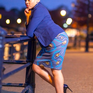 May include: A woman wearing a blue blazer and a patterned skirt stands by a black metal railing. She is wearing black high heels and has blonde hair.