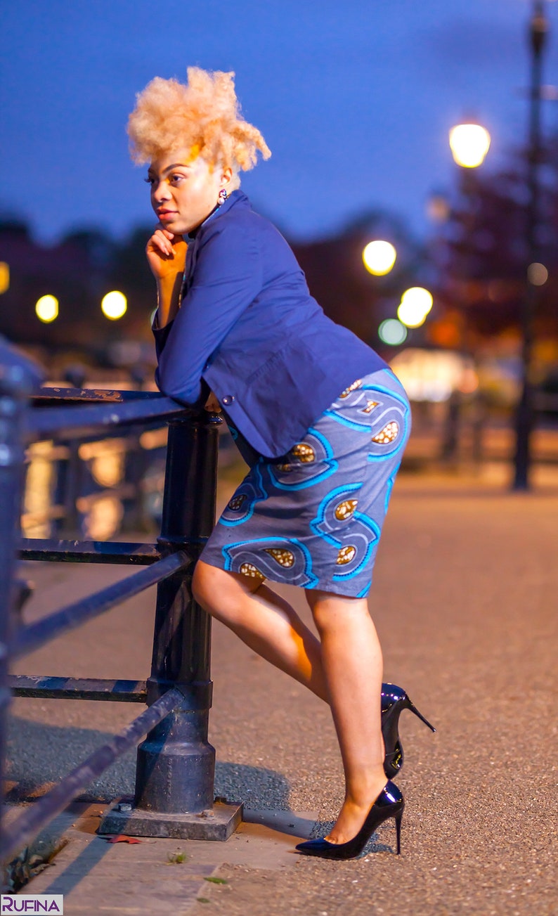 May include: A woman wearing a blue blazer and a patterned skirt stands by a black metal railing. She is wearing black high heels and has blonde hair.
