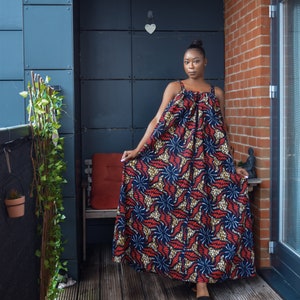 Pu&ograve; includere: Una donna indossa un lungo abito maxi fluido con una stampa geometrica colorata. L'abito ha una scollatura arricciata e una vestibilit&agrave; ampia e comoda. La donna &egrave; in piedi su un ponte di legno con un muro di mattoni e una pianta in vaso sullo sfondo.