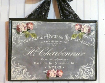 May include: A rectangular sign with a black frame and a decorative black bow. The sign has a grey background with floral and scroll designs, and text in French. The sign reads "Parfumerie D'Hygiene Rationnelle" and "H. Charbonnier".