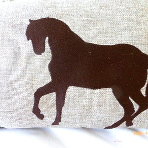 Puede incluir: Un coj&iacute;n decorativo con una silueta de caballo marr&oacute;n sobre un fondo beige. El caballo se representa en una pose de carrera, con las patas extendidas. El coj&iacute;n tiene un aspecto texturizado, lo que sugiere lino o una tela similar.