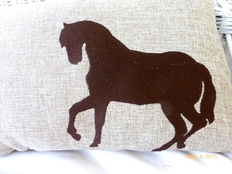 Puede incluir: Un coj&iacute;n decorativo con una silueta de caballo marr&oacute;n sobre un fondo beige. El caballo se representa en una pose de carrera, con las patas extendidas. El coj&iacute;n tiene un aspecto texturizado, lo que sugiere lino o una tela similar.