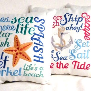 White decorative pillows with embroidered nautical themes. One pillow features a starfish and the words "sea," "shore," "life," and "reef." Another pillow displays an anchor and the phrases "Ship ahoy!" and "Sea Sail." The third pillow has a seashell.
