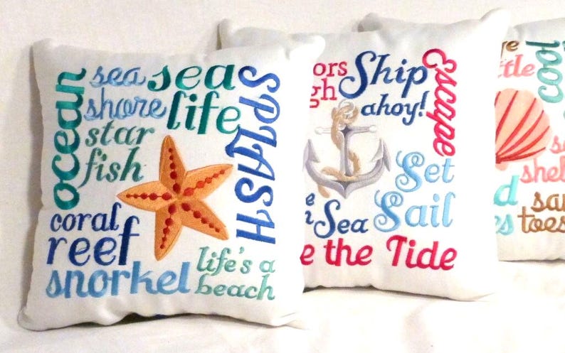 White decorative pillows with embroidered nautical themes. One pillow features a starfish and the words "sea," "shore," "life," and "reef." Another pillow displays an anchor and the phrases "Ship ahoy!" and "Sea Sail." The third pillow has a seashell.