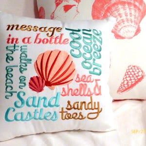A White square pillow with embroidered text and seashell designs. The pillow features phrases like "message in a bottle," "cool ocean breeze," and "sand castles." The seashells are coral and the text is in various colors.  A perfect pillow to add to your coastal decor.