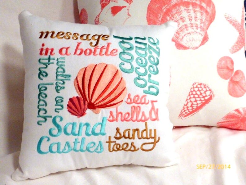 A White square pillow with embroidered text and seashell designs. The pillow features phrases like "message in a bottle," "cool ocean breeze," and "sand castles." The seashells are coral and the text is in various colors.  A perfect pillow to add to your coastal decor.