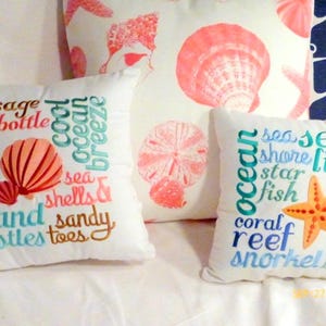 Coastal decor pillows, in the front are 2 embroidered Beach themed pillows one with a coral shell in the center and Beach themed sayings and the second has a starfish and Beach themed sayings embroidered in blue.  In the back is a ivory pillow with coral shells and starfish printed on the fabric.