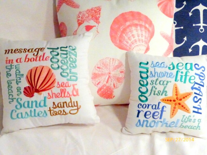 Coastal decor pillows, in the front are 2 embroidered Beach themed pillows one with a coral shell in the center and Beach themed sayings and the second has a starfish and Beach themed sayings embroidered in blue.  In the back is a ivory pillow with coral shells and starfish printed on the fabric.
