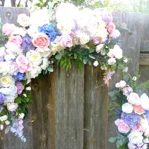 May include: A floral arrangement featuring a variety of pastel-colored flowers, including roses and hydrangeas, against a weathered wooden fence. The arrangement includes white, pink, blue, and purple blooms, with green foliage accents. A smaller arrangement is also visible.