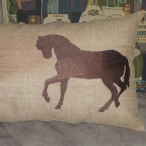 Puede incluir: Un coj&iacute;n rectangular beige con una silueta de caballo marr&oacute;n. El caballo est&aacute; representado al trote, con las patas extendidas. El coj&iacute;n tiene un aspecto texturizado, lo que sugiere una tela de yute o lino. Es un art&iacute;culo decorativo.