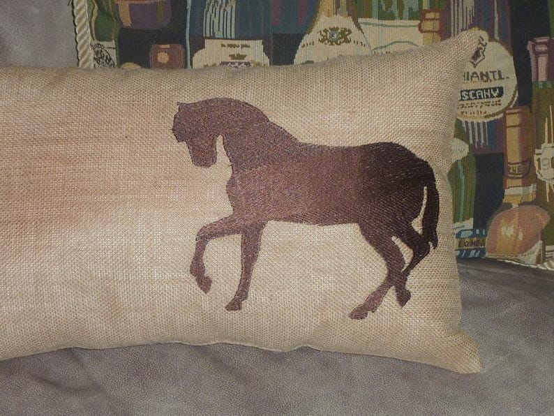 Puede incluir: Un coj&iacute;n rectangular beige con una silueta de caballo marr&oacute;n. El caballo est&aacute; representado al trote, con las patas extendidas. El coj&iacute;n tiene un aspecto texturizado, lo que sugiere una tela de yute o lino. Es un art&iacute;culo decorativo.