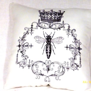 A 12x12 white Queen Bee pillow with a  fancy frame with a crown at the top and a bee in the center all printed in black.
