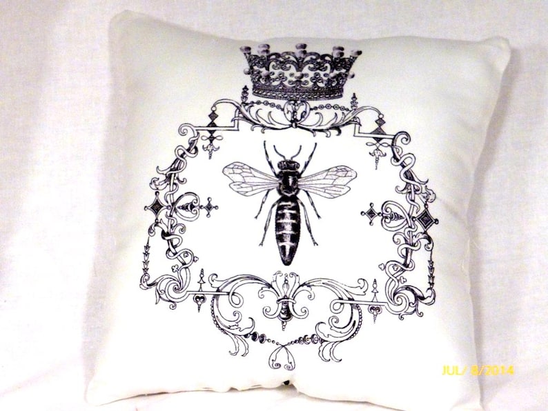 A 12x12 white Queen Bee pillow with a  fancy frame with a crown at the top and a bee in the center all printed in black.