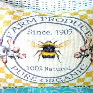 A rectangle pillow that has a yellow and off whit checked background and in the center it has a gray oval frame and pink flowers with FARM PROCUCE PURE ORGANIC printed inside the frame and a large honey bee and above the bee it reads, Since 1905 and below the bee is 100% Natural also printed in gray.  Inside the frame the background is off white.  Printed along the bottom of the pillow it has Apple Blossom Honey printed in black.