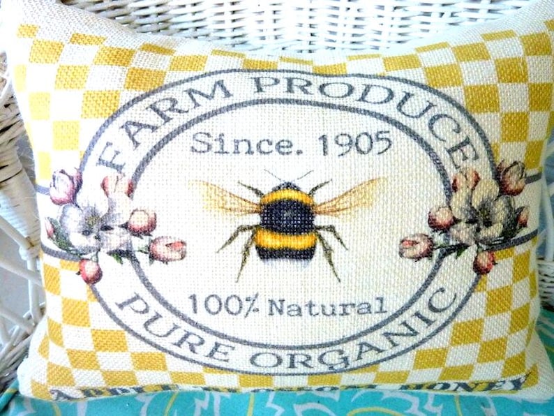A rectangle pillow that has a yellow and off whit checked background and in the center it has a gray oval frame and pink flowers with FARM PROCUCE PURE ORGANIC printed inside the frame and a large honey bee and above the bee it reads, Since 1905 and below the bee is 100% Natural also printed in gray.  Inside the frame the background is off white.  Printed along the bottom of the pillow it has Apple Blossom Honey printed in black.