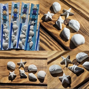 May include: A set of white plaster seashell and starfish figurines for painting, accompanied by small paint pots in blue, yellow, and purple, along with a blue-handled paintbrush, all packaged in clear plastic sleeves for individual craft kits.