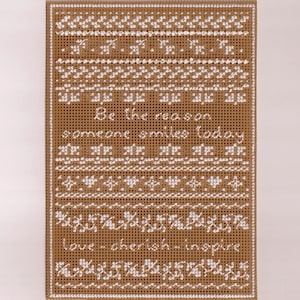 May include: A brown card with white cross-stitch embroidery. The embroidery features a pattern of hearts, stars, and flowers. The text "Be the reason someone smiles today" and "love - cherish - inspire" is embroidered on the card.