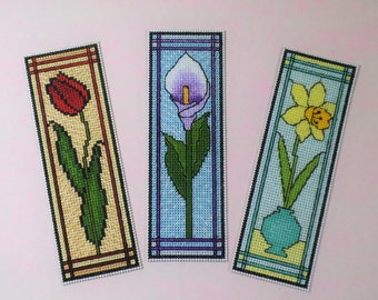 Puede incluir: Tres marcapáginas de punto de cruz con diseños florales. El primer marcapáginas tiene un tulipán rojo con hojas verdes. El segundo marcapáginas tiene un lirio de agua morado con hojas verdes. El tercer marcapáginas tiene un narciso amarillo con hojas verdes en un jarrón verde azulado.