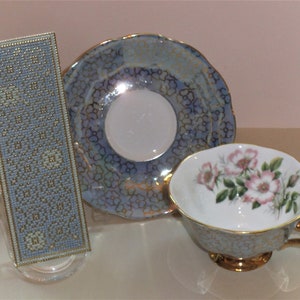 May include: A blue and gold teacup and saucer set with a floral design. The saucer has a delicate floral pattern and the teacup features a bouquet of pink roses with green leaves.