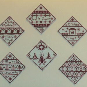 May include: Six diamond-shaped Christmas decorations with red embroidery on a white background. The decorations feature traditional Christmas themes, including "The stockings were hung", "Let it Snow", "Merry Christmas", "O Holy Night", and two geometric patterns.