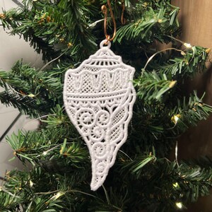 May include: White lace seashell ornament hanging on a green Christmas tree. The ornament is detailed with intricate lacework and is a delicate and elegant addition to any holiday decor.