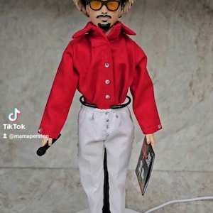 Op de afbeelding: Pop gekleed in een rood overhemd, witte broek en zwarte schoenen, met een zonnebril en een strohoed. De pop houdt een microfoon en een boek vast. Het TikTok-logo en de gebruikersnaam @mamapersten zijn zichtbaar.