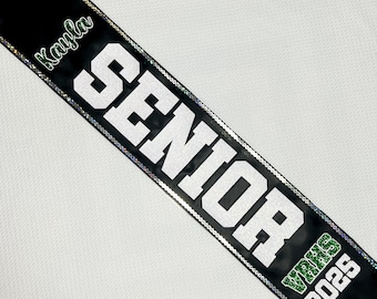 May include: Black graduation sash with the word "SENIOR" in large white letters. The name "Kayla" is in green script at the top, and "VRHS 2025" is at the bottom. The sash is trimmed with silver sequins.