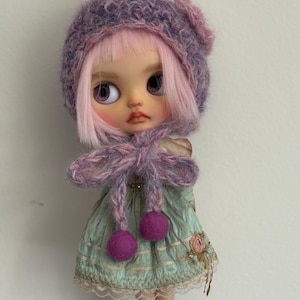 Könnte beinhalten: Eine Puppe mit rosa Haaren und großen Augen trägt eine lila Strickmütze mit einer Blume. Die Puppe trägt ein mintgrünes Kleid mit einer rosa Schleife und zwei lila Pompons. Das Outfit der Puppe hat eine Vintage-Ästhetik.